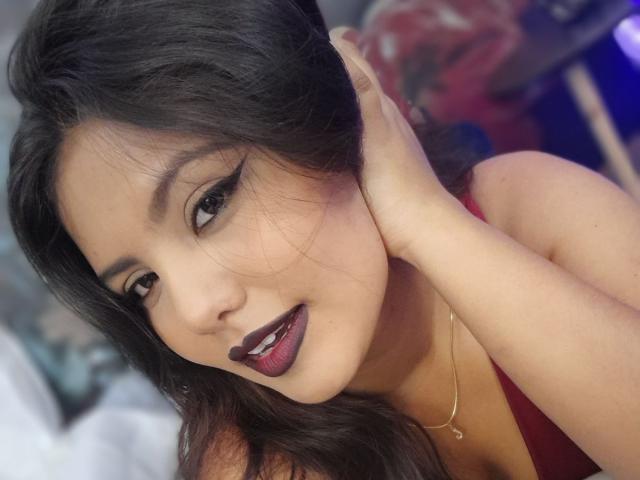 LucyDumont with dark brown hair, dark eyeliner, and purple lipstick, smiling with hand on head, wearing a red top, gold necklace, and showing off her tits.