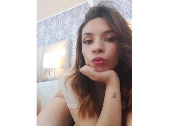 VioletaStone with brown hair, pink lips, and tattoo on right hand, pouting, in white top, resting chin on hand, in soft light, with lamp in background.