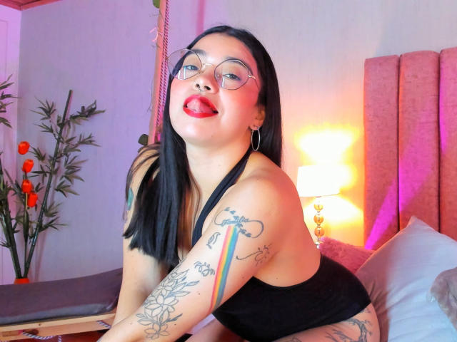 Emma Ruiz, com cabelo preto, óculos, batom vermelho, língua de fora, top preto, tatuagens nos braços, sentada na cama, cama com cabeceira rosa, abajur, flores laranjas ao fundo..