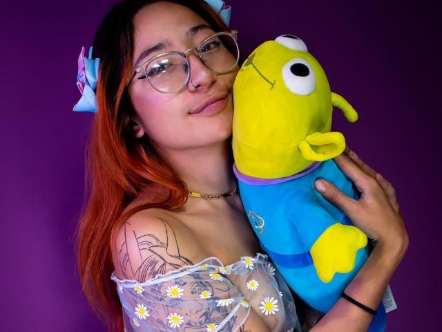 ChloeLee, avec ses cheveux roux, ses lunettes et son tatouage, tient contre sa poitrine un peluche Plimplimplim jaune, bleu et violet. Elle porte un haut transparent avec un motif de marguerites..
