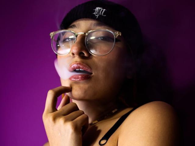 ChloeLee, à la peau foncée, portant un bonnet noir, des lunettes transparentes et un rouge à lèvres rose brillant, touchant son menton avec ses doigts, sur un fond violet..