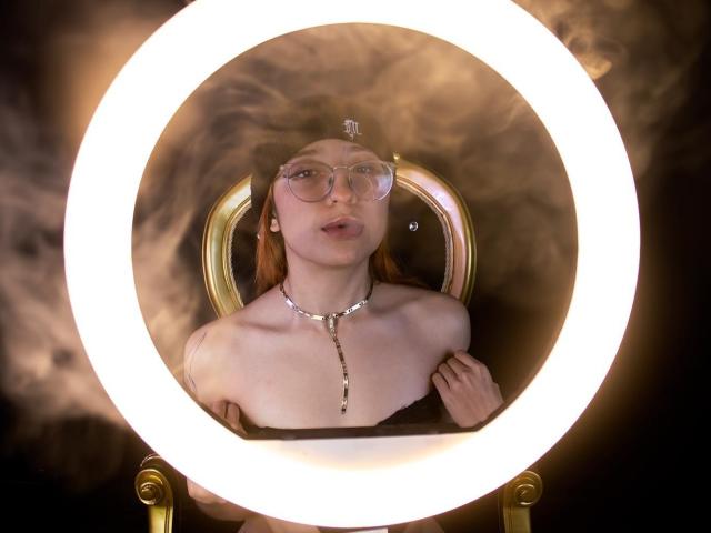 ChloeLee, avec des lunettes, une pièce de tête en forme de croix et un collier, assise sur une chaise dorée, éclairée par un anneau lumineux, montrant ses seins nus et ses cheveux roux..