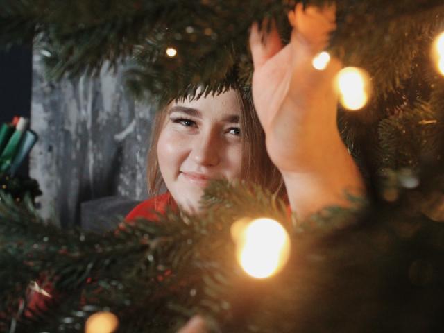 DaniellaGraceZZ's face peeking through a Christmas tree, smiling with brown eyes, red shirt, and a hand holding a branch, surrounded by glowing lights and green needles.