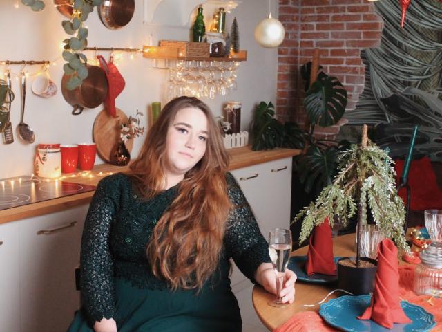 DaniellaGraceZZ con cabello rojo largo, vestido verde de encaje, sosteniendo una copa de champán, sentada en una mesa de madera con plantas, servilletas rojas y utensilios de cocina en el fondo..