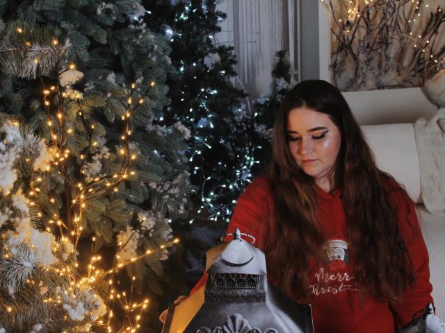 DaniellaGraceZZ, con cabello castaño largo, una camisa roja y un maquillaje sutil, sosteniendo un farol cubierto de nieve, de pie junto a un árbol de pino decorado con luces amarillas cálidas..