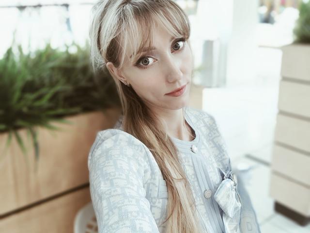 SweetyPretty has blonde hair, blue eyes, and is wearing a light gray patterned top with a small bow. She's looking at the camera with a slight smile.