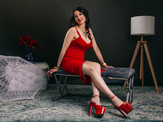 OneSpecialCerise, curvy, red dress, big tits, red heels, black wavy hair, red lipstick, sitting on bench, legs crossed, black background, white lamp, red flowers, gold jewelry.