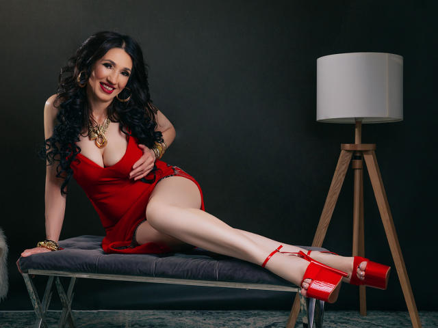 OneSpecialCerise, with big boobs, wearing a red dress, red heels, and gold jewelry, sits on a gray bench, legs crossed, smiling, next to a wooden lamp.