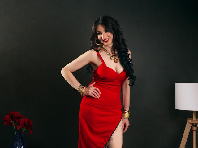 OneSpecialCerise with long black hair, red dress, cleavage, gold jewelry, smiling, hand on hip, standing, red flowers, white lamp in background. Boobs, ass, dress with thigh slit.