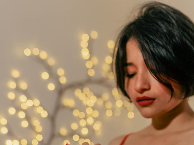 MiaLeblanc, con i capelli neri corti, rossetto rosso e occhi chiusi, indossa una maglietta rossa ed è in piedi di fronte a luci sfocate, con un'espressione dolce e serena..