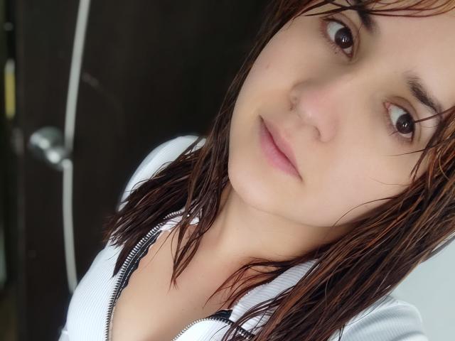 Kkatalina with wet brown hair, white zip-up, and a slight smile, looking at the camera, showing her tits slightly. Soft light, focused on her face.