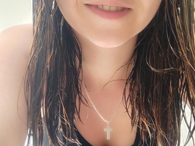 Kkatalina's wet brown hair, pale skin, and slight smile, wearing a silver cross necklace, showing her bare shoulders and black top, with her tits slightly visible.