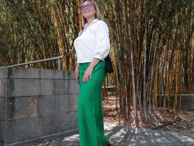 JaneTurner met een groene rok, witte blouse, zwarte schoudertas, paarse bril, lachend, staand in een bamboebos, waarbij haar achterwerk zichtbaar is met haar handen aan haar zijde..