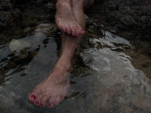 JohndaX's feet, wet with water, are crossed at the ankles, showing pink-painted toenails, with her toes slightly submerged in a rocky, shallow water source.