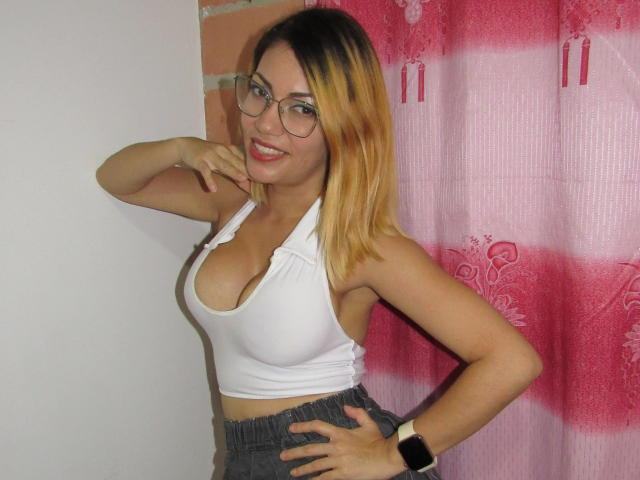 Ragazza con i capelli biondi, occhiali, top bianco che mette in mostra un seno abbondante, pantaloni grigi e uno smartwatch, sorridente, con la mano sul mento, una tenda rosa sullo sfondo..