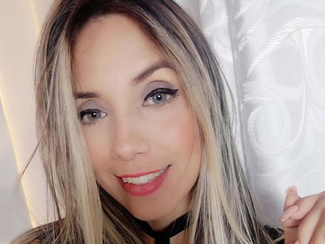 LindsaySummer heeft blond en donkerbruin haar, blauwe ogen, zwarte eyeliner, rode lipstick en een zwarte choker, en ze glimlacht met licht zichtbare tanden..