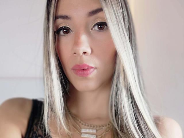 LindsaySummer ha capelli biondi e neri, occhi marroni, labbra rosa e indossa una maglia nera in pizzo con una collana dorata, guardando direttamente la fotocamera con un'espressione neutra..