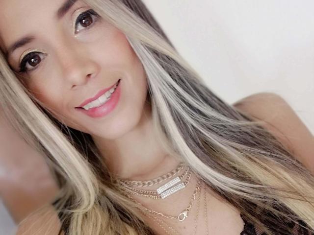 Lindsay, zomer. Lang blond en bruin haar, lachend met roze lippen, bruine ogen, draagt gouden kettingen en een zwarte top, waarbij haar borsten lichtjes in beeld zijn..