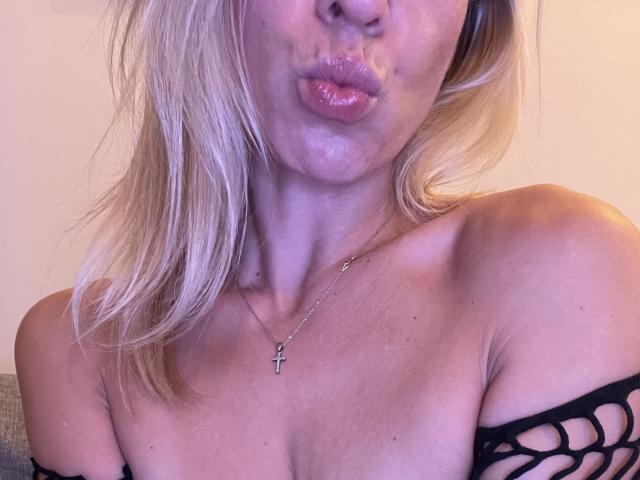 SandraSwweet, con labbra rosa carnose, capelli biondi, una croce al collo e spalle scoperte, indossa una maglia nera a rete che mette in mostra i suoi piccoli seni..