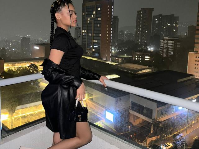 MilaZarate with black braided hair, wearing a black dress and leather jacket, standing on a balcony at night, holding a black purse, city lights in the background.