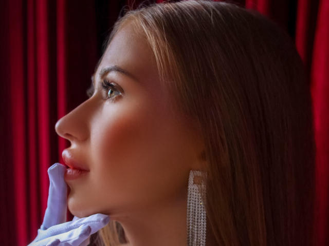 Das Profil von Violetta Steward, mit weißen Handschuhen und langen Diamant-Ohrringen, mit roten Lippen und braunen Haaren, die zur Seite blickt. Ihr Hintern ist nicht sichtbar..