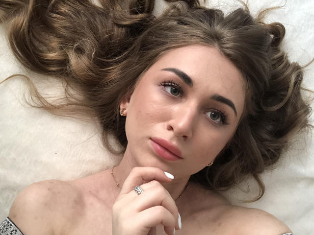 El rostro de BeataAngel, con cabello castaño ondulado, ojos verdes, labios rosados, uñas blancas, aretes dorados y un anillo, sobre una superficie blanca, mirando a la cámara con una ligera sonrisa..