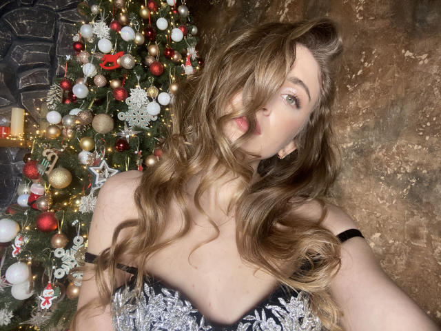 BeataAngel with long, wavy brown hair, tongue out, wearing a sparkly black dress, standing in front of a Christmas tree with red, gold, and white ornaments.