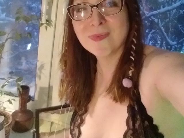 IrunchaArt has pale skin, brown hair in braids with pink beads, glasses, and is wearing a lace black top revealing her small tits. She's smiling at the camera.