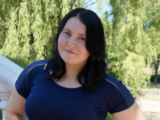 LorriMindy, eine etwas korpulente Frau mit schwarzen, schulterlangen Haaren, trägt ein dunkelblaues Hemd und zeigt ihre großen Brüste. Sie steht draußen, im Hintergrund sind grüne Bäume zu sehen..