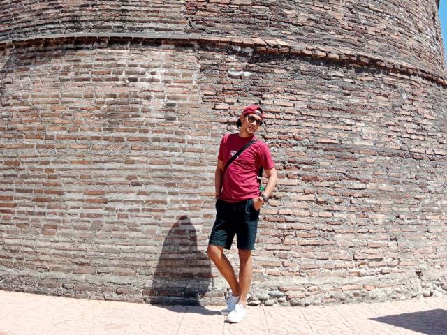 Jhe69X se tient devant un mur de briques, portant une casquette rouge, un t-shirt rouge, un short noir, des chaussettes blanches et des chaussures blanches, avec une ombre au sol..