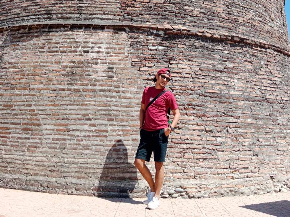 Jhe69X steht vor einer Ziegelwand, trägt eine rote Mütze, ein rotes T-Shirt, schwarze Shorts, weiße Socken und weiße Schuhe, und es wirft ein Schatten auf den Boden..