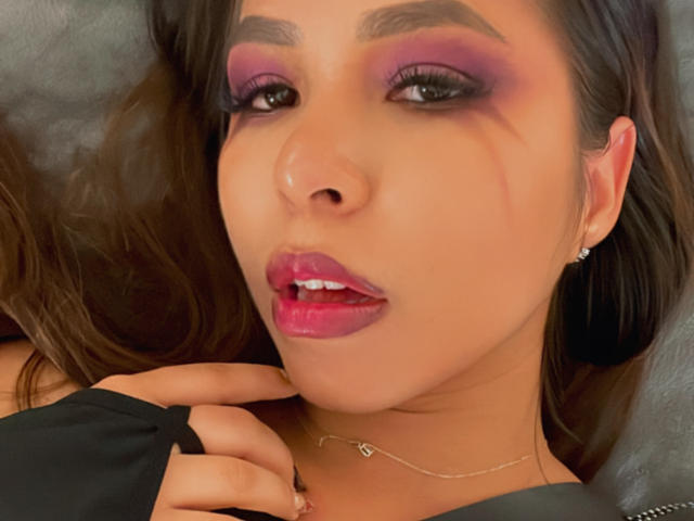 MeganWisse has purple eyeshadow, red lipstick, and a black fingerless glove. Her hand touches her chin. She has a silver necklace and dark brown hair.