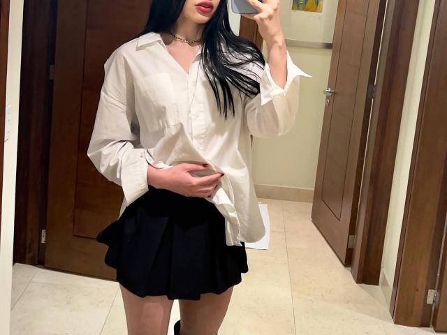 LolaHeaven, con lunghi capelli neri, indossa una camicia bianca ampia e una gonna nera, tiene il telefono per un selfie allo specchio, mostrando una mano sulla pancia, rossetto rosso, porte di legno, pavimento piastrellato..