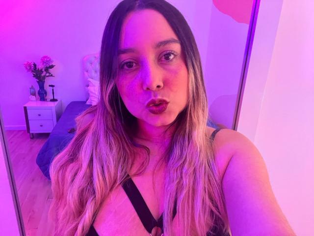 AmayaJhons met roze en paarse verlichting, vollere lippen, lang golvend blond haar, gekleed in een zwarte haltertop, waarbij haar grote borsten zichtbaar zijn, houdt een camera vast voor een selfie in de slaapkamer..