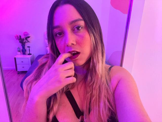 AmayaJhons avec un éclairage rose et violet, doigt dans la bouche, longs cheveux blonds et bruns, robe noire à bretelles, regardant la caméra, montrant ses seins, expression sexy, sol en bois, miroir..