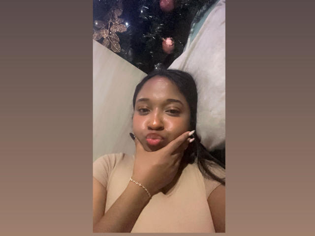 MelanyScoth with brown skin, pouty lips, and black hair, touching her cheek with her hand, wearing a beige top, and a gold bracelet, looking slightly bored.