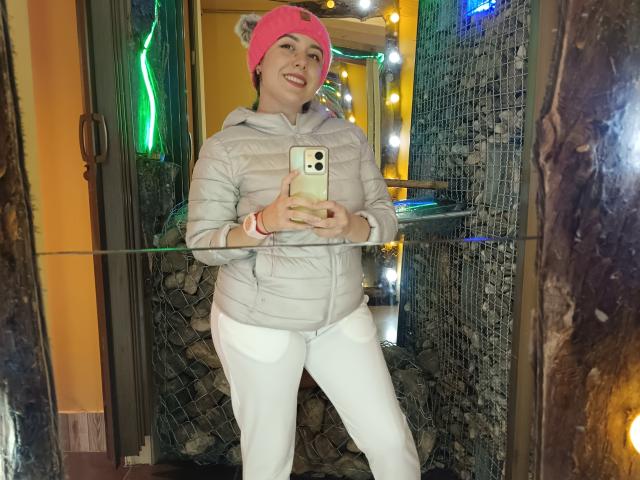 MabyLegrand with pink hat, grey puffy jacket, white pants, holding phone, smiling, standing in front of mirror, reflecting her ass and phone. Reflective wall and lights behind.