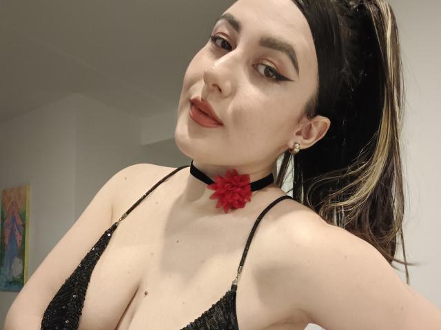 MabyLegrand with dark hair in a ponytail, black choker with red flower, black lace bra, and eyeliner, smiling at camera, showing her tits.