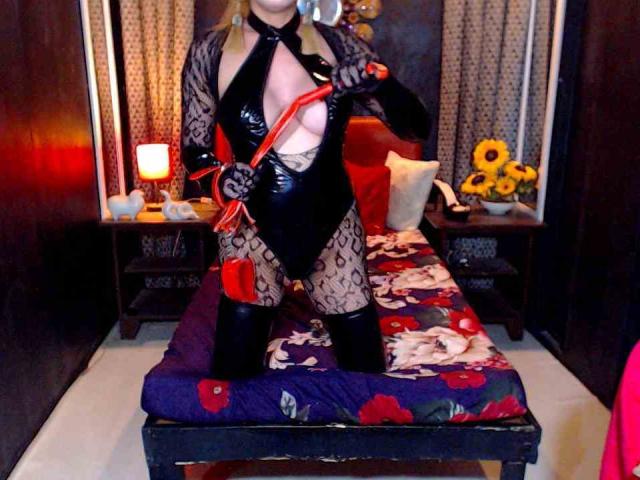 SaintHornyTs in a black, shiny, latex, open-cleavage, fishnet, bodysuit, holding a red leather flogger, standing on a bed with a floral, purple, red, and white bedspread.