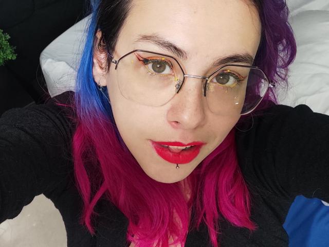 NatashaLau with purple and blue hair, red lipstick, glasses, nose ring, and lip piercing, wearing a black shirt, looking at the camera with a slight smile.
