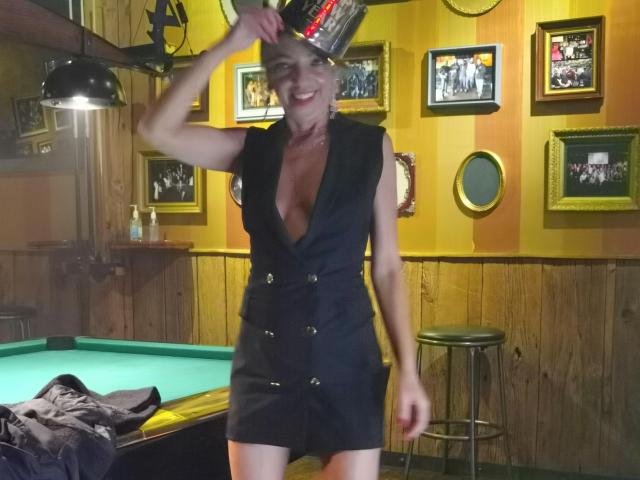 LunaBriella, con senos pequeños, vestida con un vestido negro sin mangas y escote pronunciado, sosteniendo una lata de cerveza sobre su cabeza, sonriendo en un bar con poca luz, de pie junto a una mesa de billar..