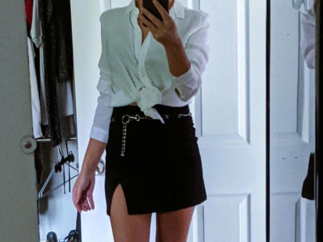 LunaBriella indossa una camicia bianca annodata, una minigonna nera con una fessura e una cintura a catena, e tiene un telefono davanti a uno specchio, mostrando le sue gambe e il suo sedere scoperti..