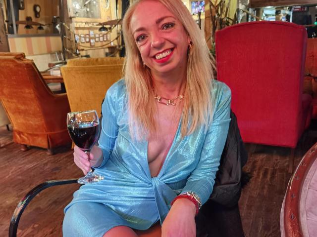 LunaBriella with blonde hair, smiling, wearing a shiny blue dress with deep cleavage, holding a glass of red wine, sitting in a cozy room with red and orange chairs.