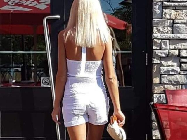 LunaBriella has blonde hair, a white strapless romper, and is holding a white purse. Her ass is small and her back is to the camera.
