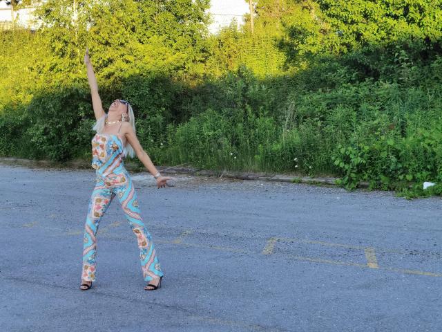 LunaBriella, con cabello rubio, vestida con un traje de baño ajustado, de una sola pieza y con estampados florales coloridos, y sandalias negras, bailando con los brazos levantados en una carretera de asfalto gris, rodeada de vegetación..