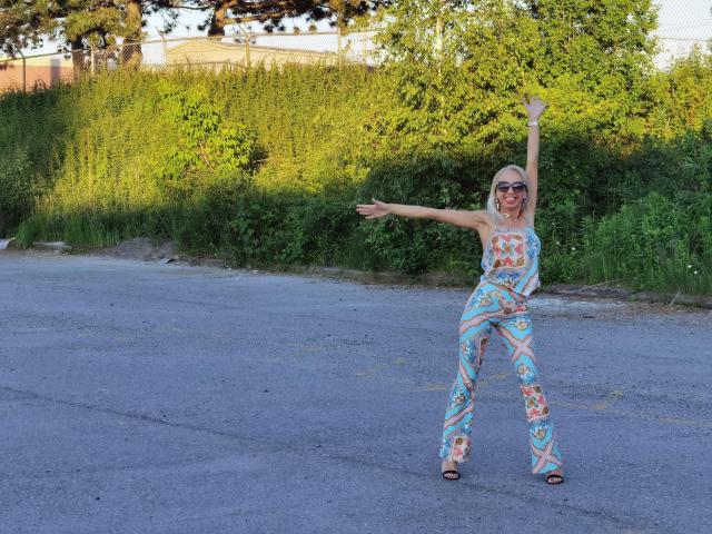 LunaBriella with blonde hair, wearing colorful patterned pants and top, raising right arm, pointing with left, standing on empty road, wearing sunglasses, smiling, in daytime.