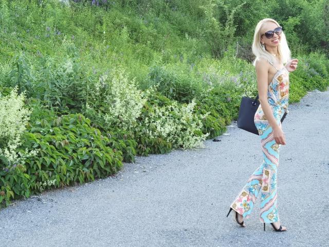 LunaBriella met blond haar, gekleed in een kleurrijke, gestreepte, mouwloze jumpsuit, zwarte hakken en een zonnebril, loopt over een grindpad en houdt een zwarte clutch vast..