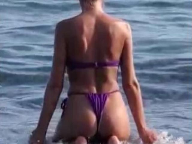 LunaBriella está arrodillada en el agua, de espaldas, vistiendo un bikini de hilo púrpura que muestra su trasero y sus pequeños pechos. Su espalda está bronceada y está a la luz del sol..