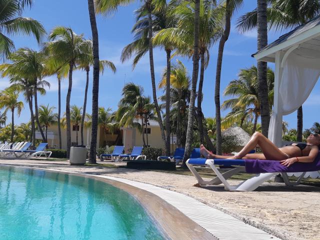 LunaBriella, ein attraktives Mädchen mit gebräunter Haut, entspannt sich auf einer blauen Liege am Pool, trägt einen schwarzen Bikini, wobei ihr Hintern und ihre Brüste freiliegen, unter Palmen..