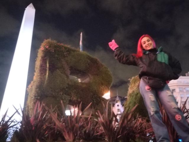 AmyStreetwise69 with bright red hair, black jacket, green scarf, and ripped jeans, raising pink-gloved hand, standing near large moss-covered letters, under dark night sky.